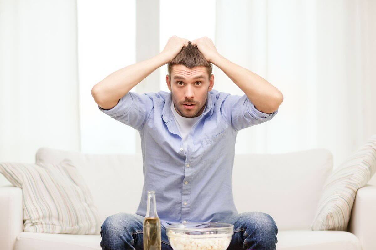 Uomo con mani nei capelli guarda TV, pop corn, birra